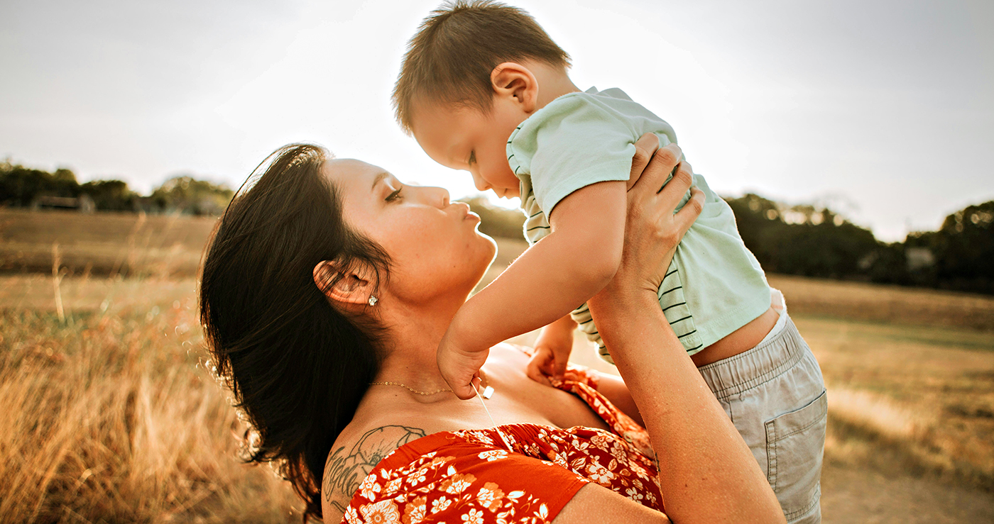 A mother in red dress holds an infant baby above her shoulders and mimes a kiss.