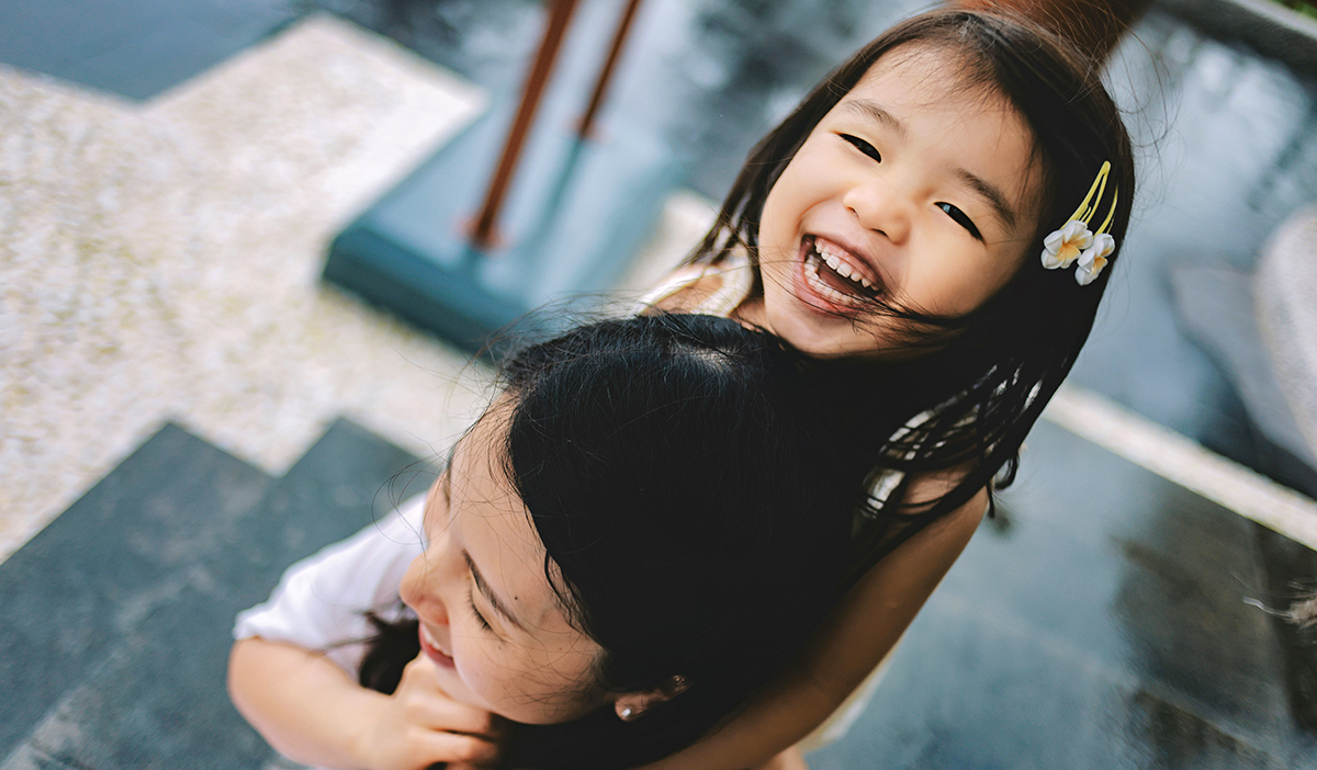 Two young Vietnamese girls hug viewed from above with one girl turns head upward toward the camera while laughing.