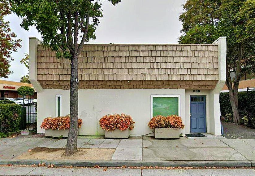 New offices of DrsHernandez.com, small beige building with wood shake roof, blue door, orange flowers in front, tall trees on right side.