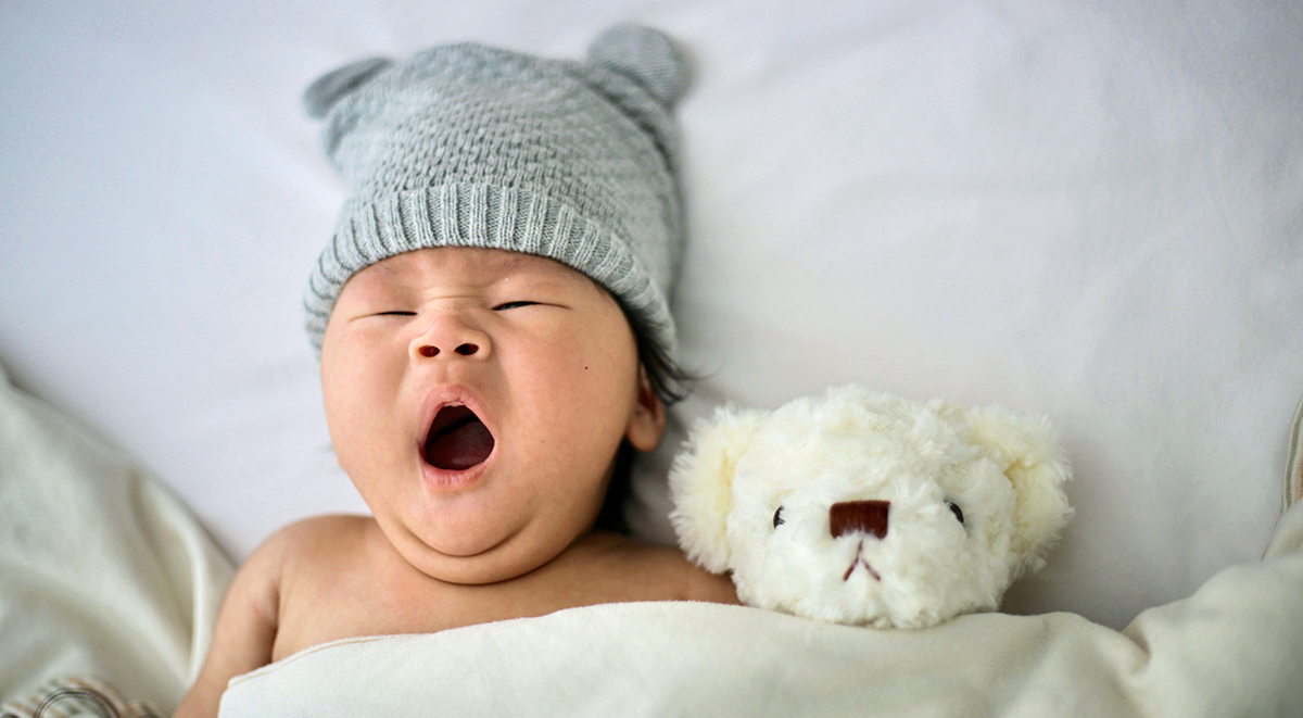 Chinese baby lying on back asleep with mouth open next to teddy bear and wearing gray knitted cap