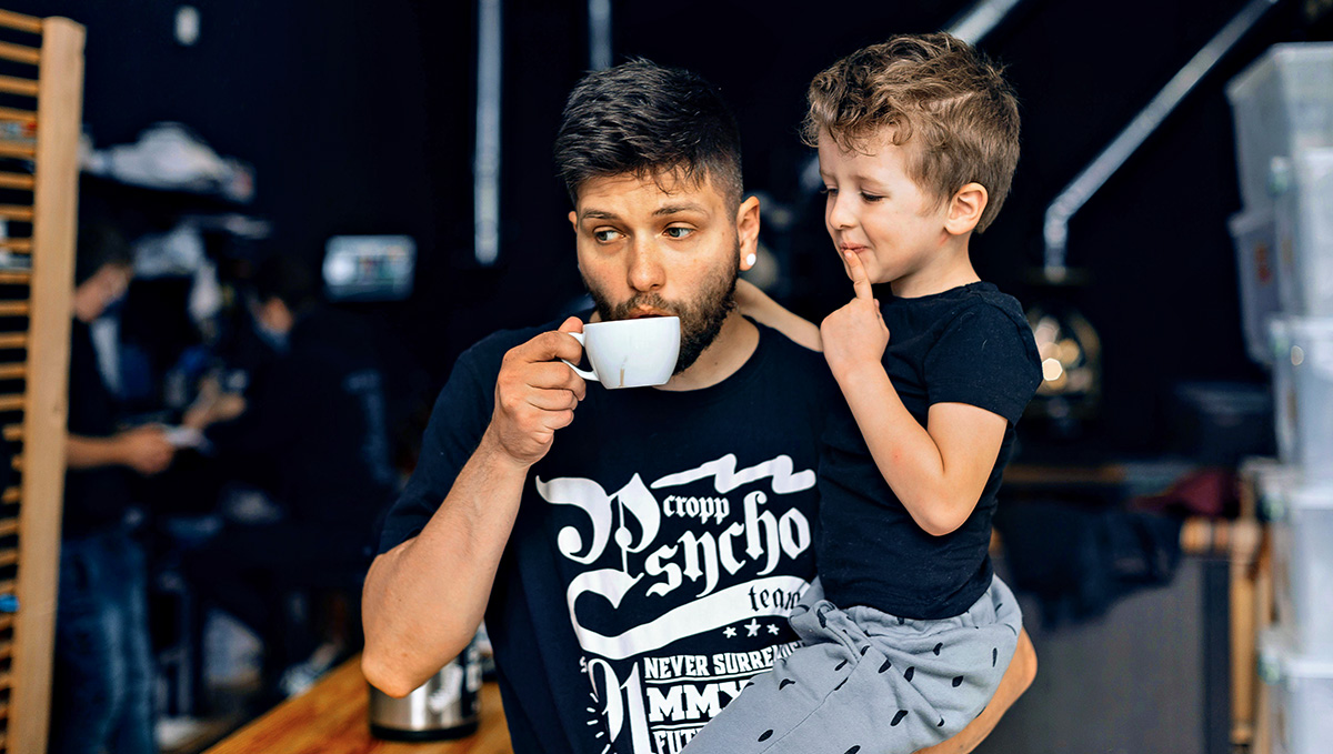 Father drinks coffee while leaning with right elbow on wood grain table with son in left arm smiling.
