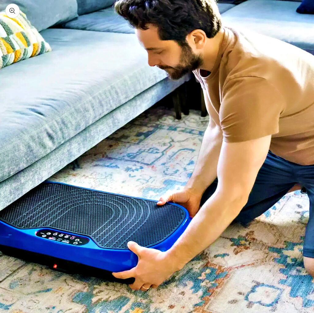 Young man with beard slides a blue vibration plate under a bed.