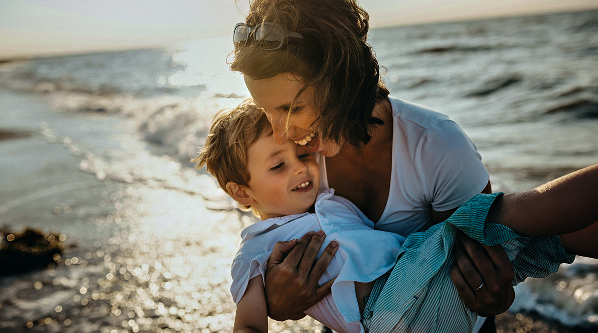 Smiling mother holds laughing baby boy in arms with sunlit ocean in background.